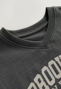 Donkergrijze katoenen T-shirt met een V-hals, geribbelde kraag en witte grafische print. De textuur lijkt zacht en licht geborsteld.