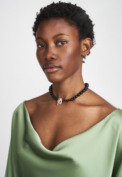Μαύρο κολιέ με χάντρες και ασημένια λεπτομέρεια, φορεμένο στον γυμνό ώμο. Το μοντέλο φοράει ένα πράσινο draped τοπ, τονίζοντας το ντεκολτέ και τα κοσμήματα.