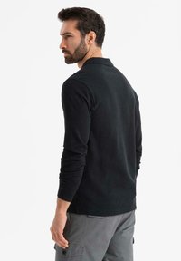 Man med mörkt hår och skägg som bär en långärmad svart polo och grå cargobyxor, tittande åt vänster mot en vit bakgrund.