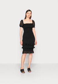 Vestido preto com mangas curtas em tule bufante, corpete ajustado e bainha em camadas com babados; tecido suave com textura franzida por todo o lado. Mule preta de salto alto.