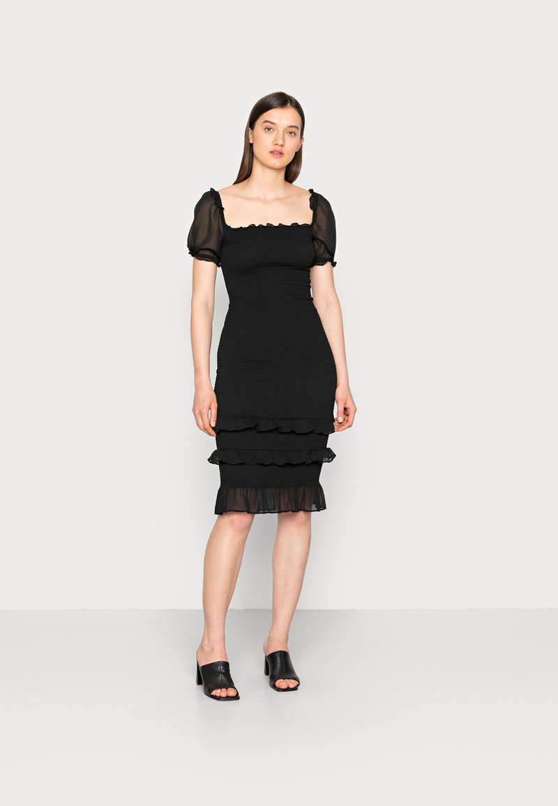 Vestido preto com mangas curtas em tule bufante, corpete ajustado e bainha em camadas com babados; tecido suave com textura franzida por todo o lado. Mule preta de salto alto.