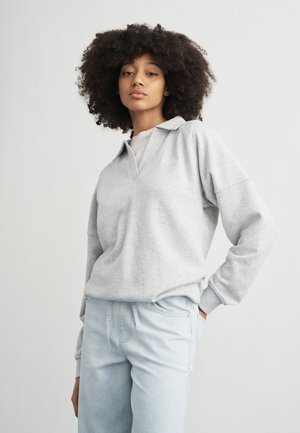 Νεαρή γυναίκα με σγουρά μαλλιά, φοράει ανοιχτό γκρι πουλόβερ και ανοιχτό μπλε τζιν, στέκεται με το ένα χέρι στην τσέπη μπροστά σε απλό φόντο.