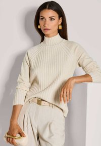 Crème gebreide trui met een hoge kraag, met een textuurpatroon. Gecombineerd met lichtgekleurde broeken en een neutrale clutch.