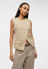 Bēšas bezpiedurkņu veste ar V veida kakla izgriezumu, priekšējo drapējumu un sudraba akcentu. Satur divas priekšējās kabatas un ir kombinēta ar atbilstošām platām biksēm.