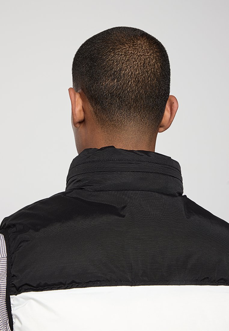 Homme aux cheveux courts portant une veste noire et blanche, vu de dos sur un fond gris uni.