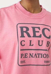 Růžová bavlněná mikina s kulatým výstřihem, zdobená tmavě šedým grafickým potiskem, který obsahuje "REC CLUB," "P.E NATION" a "EST. 1980."
