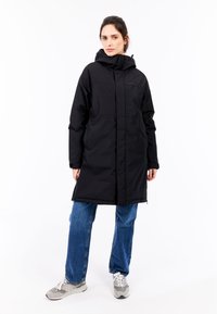 Lang sort parka med høj krave og hætte, der har lynlåslukning og sidelommer, sammen med blå cowboybukser og grå sneakers.