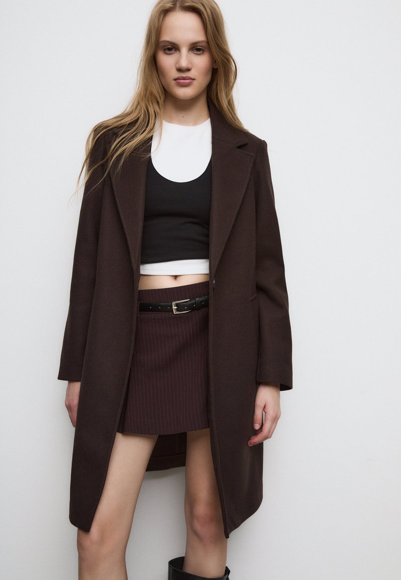 Bruine wollen jas met klaprokken over een zwarte crop top en een minirok met streepjes, aangevuld met een zwarte riem en kniehoogde laarzen.