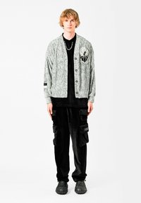 Cárdigan gris texturizado con botones negros, superpuesto a una camisa negra. Pantalones cargo de pana negros con bolsillos laterales y zapatos negros texturizados.