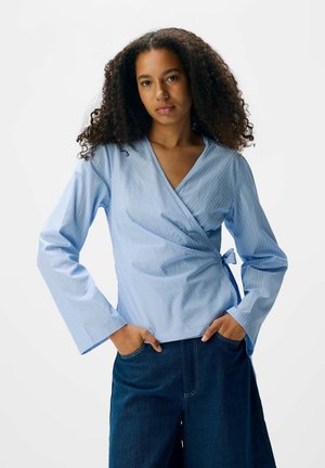 Chemise bleu clair enroulée avec des rayures verticales blanches, décolleté en V, manches longues et détail de nœud sur le côté, associée à des culottes en denim foncé.