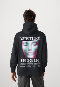 Sweatshirt preto com um design gráfico apresentando um rosto estilizado em verde-água e rosa, e o texto "VERTIERE BERLIN" nas costas e na manga.