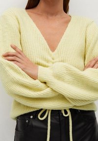 Pull jaune clair tricoté avec un col en V profond, une texture côtelée et des manches surdimensionnées, associé à un pantalon en cuir noir avec un détail noué à la taille.