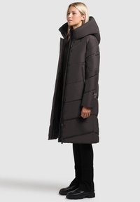 Kvinna i svart lång pufferjacka med hög krage och huva, svarta byxor och svarta stövlar, stående i profil mot en grå bakgrund.