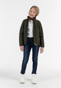 Giacca softshell verde oliva con colletto alto, chiusura con zip e polsini elasticizzati, indossata sopra una t-shirt bianca e jeans blu. Sneakers bianche.