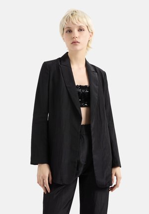 Blazer nero in tessuto stropicciato, caratterizzato da un design aperto, maniche lunghe e una vestibilità comoda, abbinato a un completo nero coordinato.