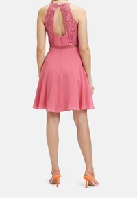 Pink dress with a floral lace bodice, halter neck design, and a keyhole back. Flared skirt with a smooth texture. Worn with orange heels.