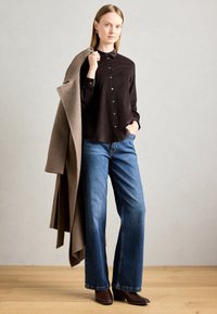 Woman wearing black buttoned shirt, blue wide-leg jeans, and brown boots, holding a long brown coat over her shoulder, standing on wooden floor.