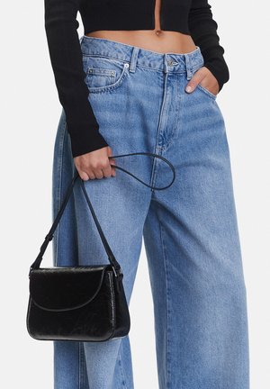 Persona che indossa jeans blu larghi a vita alta e maglione nero corto, con una borsa a tracolla in pelle nera tenuta in una mano.