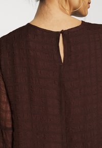 Femme montrant le dos d'un chemisier marron foncé texturé avec une fermeture à bouton en forme de trou de serrure et une grande boucle d'oreille en or visible sur le côté.
