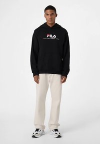 Felpa nera con cappuccio regolabile con coulisse, polsini e orlo a coste, con logo testuale, abbinata a pantaloni beige chiaro e sneakers bianche.