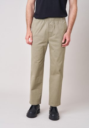 Man die beige elastische taillebroeken, zwarte schoenen en een zwart overhemd draagt, staand tegen een effen lichte achtergrond.