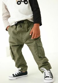 Pantaloni cargo verde oliva con tasche laterali, vita elastica e polsini alle caviglie, abbinati a sneakers nere con suole e lacci bianchi.