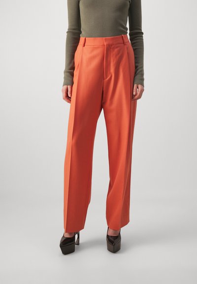 Pantaloni portocalii cu talie înaltă, cu pliuri, croi ajustat și o textură netedă, asortați cu pantofi cu platformă maro.