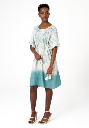 Kobieta w białej i morsko-zielonej sukience w wzory paisley sięgającej do kolan, z paskiem z frędzlami oraz beżowych sandałach na wysokim obcasie, stojąca na białym tle.