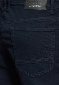 Marineblaues strukturiertes Gewebe einer Hose, das eine Gesäßtasche und ein rechteckiges Etikett zeigt, das am Bund genäht ist.