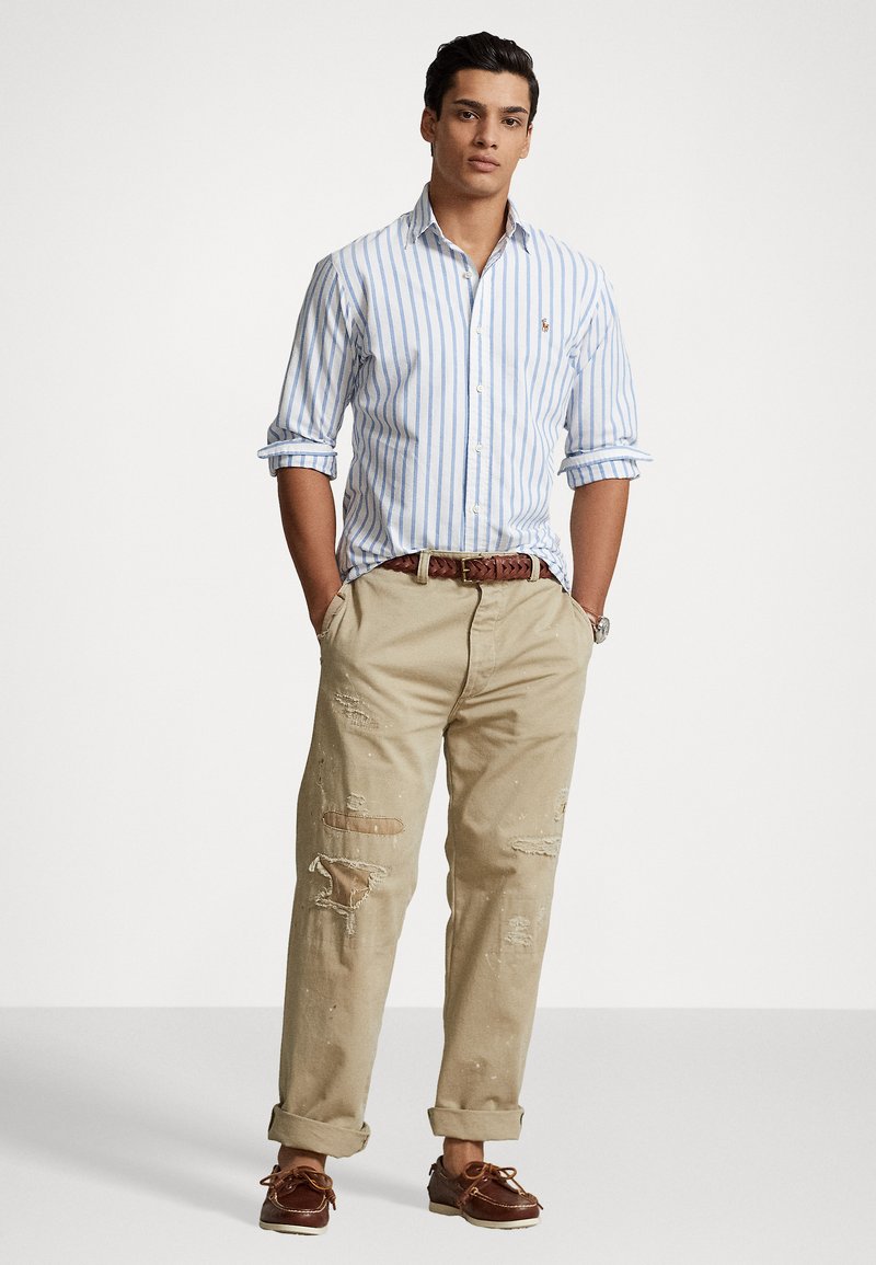 Camicia a righe blu chiaro e bianca con bottoni; pantaloni beige usurati con strappi; cintura intrecciata marrone; scarpe da barca marroni.