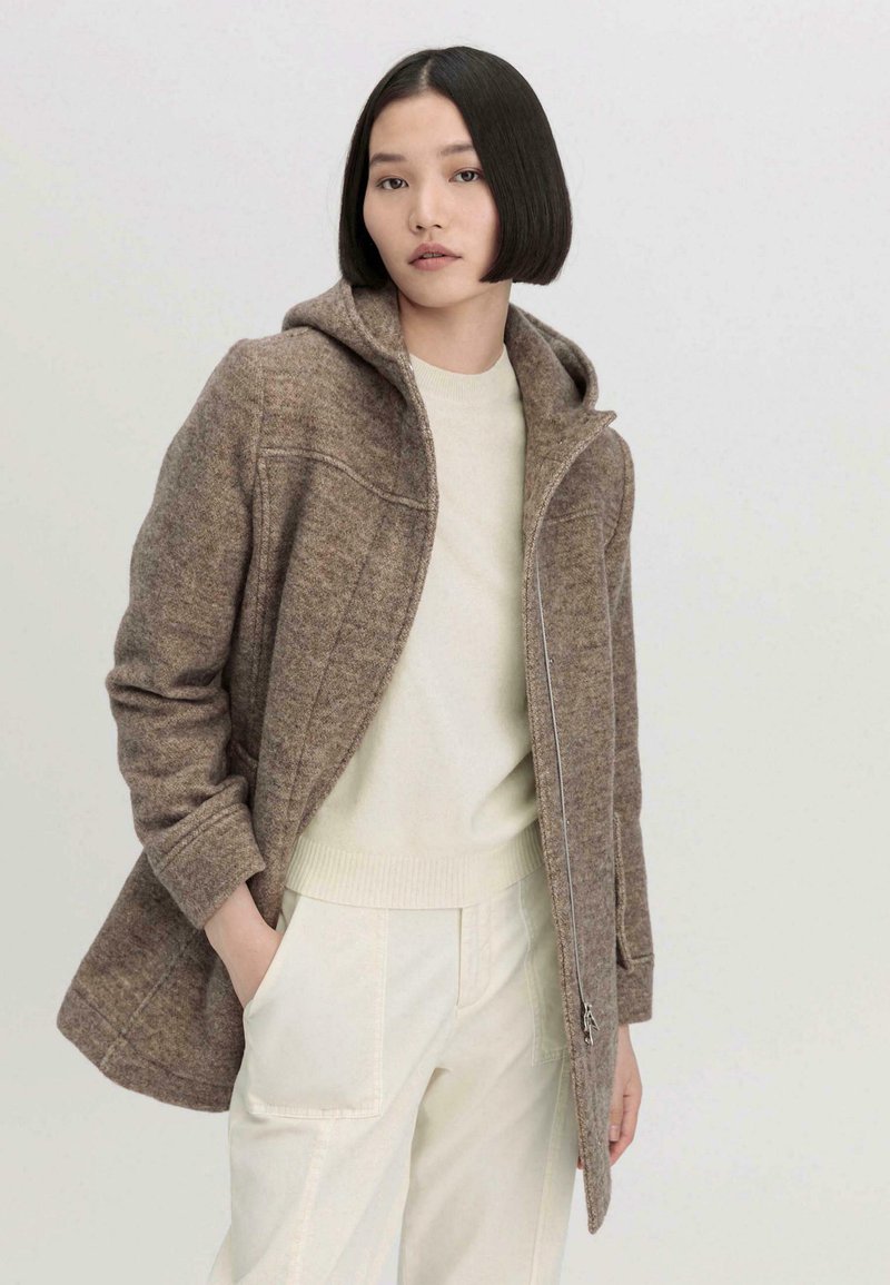 Bruine wollen jas met een capuchon, met een ritssluiting en twee zijzakken. Gecombineerd met een crèmekleurige trui en lichtgekleurde broeken.