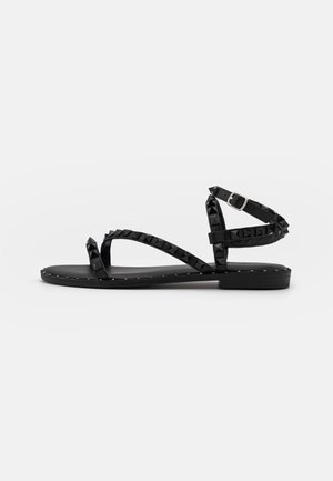 Sort flad sandal med nittebesatte remme, ankelspænde og syningsdetalje på sålen, set fra siden mod en hvid baggrund.