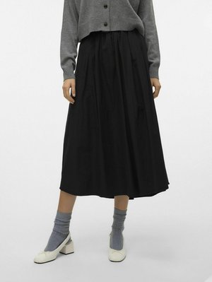 Jupe noire, longueur cheville avec une taille froncée. Portée avec un pull gris, des chaussettes grises et des chaussures blanches à talon carré.