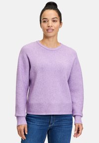 Lila gerippter Pullover mit langen Ärmeln und Rundhalsausschnitt, mit strukturierter Oberfläche und leicht lockerer Passform. Kombiniert mit blauen Jeans.
