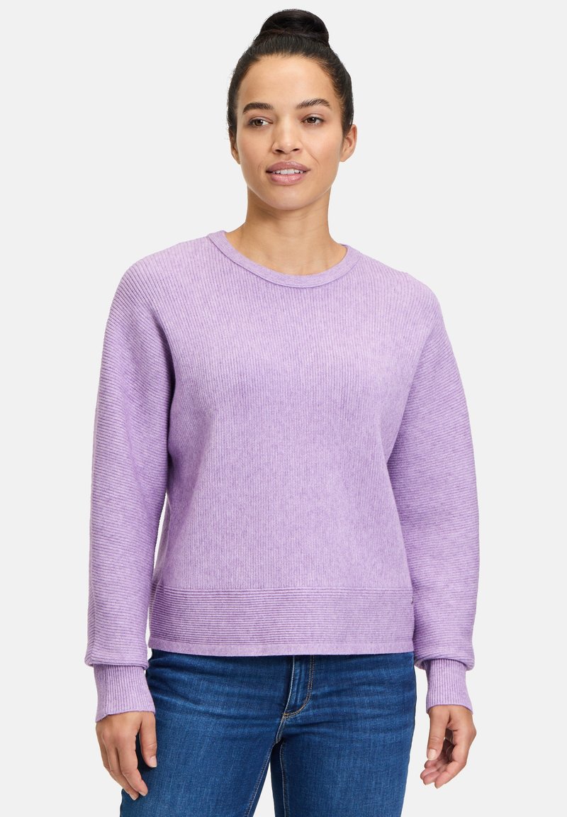 Lila gerippter Pullover mit langen Ärmeln und Rundhalsausschnitt, mit strukturierter Oberfläche und leicht lockerer Passform. Kombiniert mit blauen Jeans.