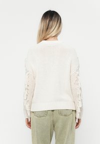 Strickpullover in Creme mit fransigen Ärmeln und geripptem Saum. Weiche Textur und lockere Passform, kombiniert mit hellgrünen Jeans.