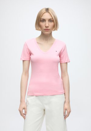 Femme aux cheveux blonds courts portant un t-shirt rose à col en V et manches courtes, ainsi qu'un pantalon blanc, debout devant un fond clair uni.
