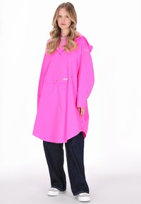 Rózsaszín vízálló esőköpeny kapucnival, elülső zsebbel és gombokkal. Sötét farmernadrág és fehér tornacipő fölött viselve, laza, oversized szabással.