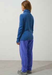 Blå, tjock stickad tröja med hög krage, kombinerad med ljuslila, löst sittande byxor. Bakgrunden är neutral och minimalistisk.