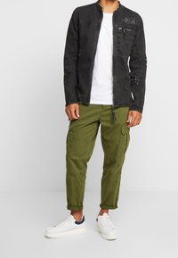 Mies, joka pitää yllään mustaa kulutettua vetoketjullista takkia, valkoista t-paitaa, oliivinvihreitä cargo-housuja käännetyin lahkeineen sekä valkoisia lenkkareita, joissa on tummansinisiä yksityiskohtia.