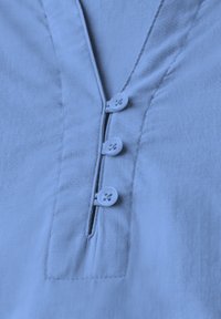 Camicia azzurra chiara con colletto a V, caratterizzata da tre bottoni e da cuciture sottili lungo la patta e i lati. Tessuto dalla consistenza liscia.