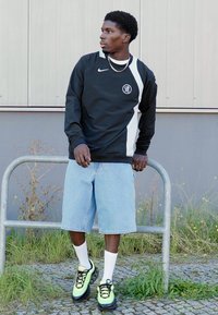 Sort langærmet Nike-top med hvide detaljer, lyseblå denim shorts, hvide sokker og neongrønne sneakers. Står nær et metalgelænder.