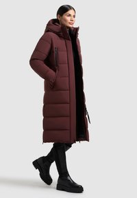 Abrigo acolchado burdeos con capucha, bolsillos con cremallera, diseño acolchado y una silueta larga, combinado con botas negras hasta la rodilla.