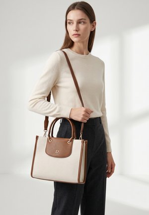 Femme portant un pull crème et un pantalon foncé, tenant un sac à main beige et marron avec une bandoulière, regardant sur le côté.