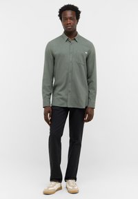 Zielona koszula z guzikami, z kieszenią na piersi, długimi rękawami i miękką teksturą, w połączeniu z ciemnymi spodniami i casualowymi trampkami.
