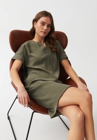 Olive green short-sleeve dress with a V-neckline, smooth texture, and a relaxed fit, worn by a seated person in a brown leather chair.