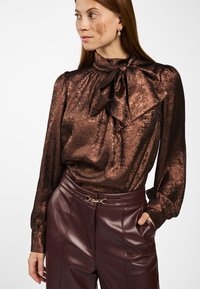 Vrouw met lang rood haar, gekleed in een glanzende bruine blouse met een grote strik en bordeauxrode leren broek, hand in de zak, kijkend naar links.