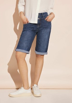 Shorts en denim avec un lavage bleu foncé, ourlets roulés et poches avant. Associés à des baskets blanches et une chemise légère à boutons.