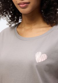 Γυναίκα που φοράει ανοιχτογκρι μπλουζάκι με στρογγυλή λαιμόκοψη, με ένα μικρό ροζ ριγέ καρδιά στο αριστερό μέρος του στήθους.