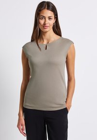 Světle olivově zelený top s krátkými rukávy a detailním průstřihem na výstřihu, vyrobený z hladkého materiálu, v kombinaci s černými kalhotami.